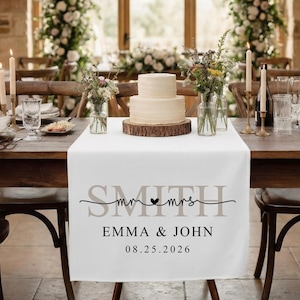 Puede incluir: Una mesa de recepción de bodas con un camino de mesa blanco con los nombres "Smith", "Emma & John" y la fecha "08.25.2026". Un pastel de dos pisos está sobre una base de madera. Arreglos florales y velas decoran la mesa.