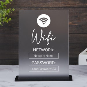 以下が含まれることがあります： 白いアクリル製の看板に、wifiのシンボルと「Wifi」という文字が筆記体で書かれています。看板には、「ネットワーク」と「パスワード」という言葉も書かれており、ネットワーク名とパスワードを入力するための空白があります。看板は黒いスタンドの上に置かれ、白い大理石の表面に置かれています。