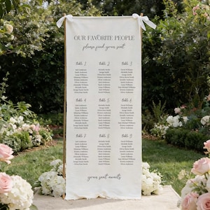 Puede incluir: Un plano de asientos de boda de color crema con el texto "OUR FAVORITE PEOPLE please find your seat." El gráfico enumera los nombres debajo de los números de mesa 1-9. El letrero se muestra al aire libre, rodeado de vegetación y flores.