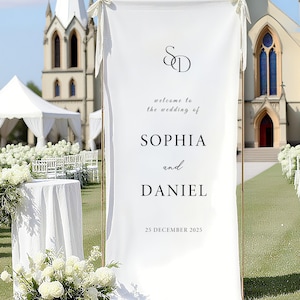 Puede incluir: Un cartel de bienvenida de boda blanco con los nombres SOPHIA y DANIEL, y la fecha 25 DE DICIEMBRE DE 2025. El cartel está enmarcado con una estructura metálica dorada y está colocado al aire libre frente a una iglesia.