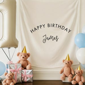 May include: A celebratory scene featuring a large number one balloon, teddy bears in party hats, and a banner reading "HAPPY BIRTHDAY James". Silver wrapped gifts with pink ribbons and blue and white balloons create a festive atmosphere.