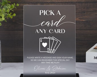 May include: A clear acrylic sign with a black stand, featuring the text "Pick a card, any card" in white. The sign also includes a graphic of playing cards and instructions for guests to write a message and sign their name. The sign is personalized with the names "Oliver & Johassa" and the date "11.12.2024".