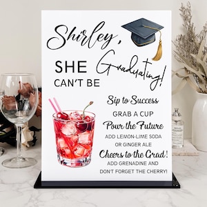 May include: A graduation-themed sign with the text "Shirley, She Can't Be Graduating!" and a cocktail illustration. The sign includes instructions for a celebratory drink, a graduation cap graphic, and is displayed on a black stand.