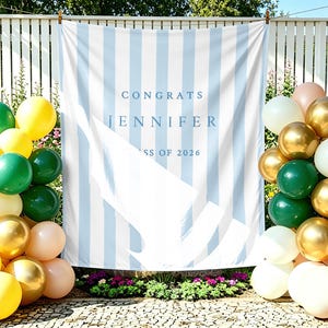 Può includere: Un banner di laurea a righe blu e bianche con la scritta "CONGRATS JENNIFER CLASS OF 2026". Palloncini dorati, bianchi, verdi, gialli e color pesca sono disposti su entrambi i lati, creando un'atmosfera festosa.