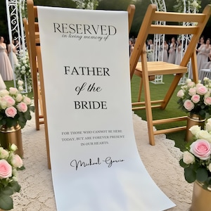 May include: A white banner with black text that reads "RESERVED in loving memory of FATHER of the BRIDE." The banner is displayed on a wooden chair at an outdoor wedding. The text "FOR THOSE WHO CANNOT BE HERE TODAY, BUT ARE FOREVER PRESENT IN OUR HEARTS. Michael Garcia" is also visible.