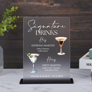 Puede incluir: Un letrero acrílico transparente con base negra muestra "Signature Drinks". Enumera "His" Espresso Martini y "Hers" Dirty Martini, con ingredientes. El letrero incluye los nombres "William & Amelia" y la fecha "10.5.2025."