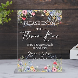 Op de afbeelding: Transparant acryl bord met bloemversieringen en de tekst "PLEASE ENJOY - THE - Flower Bar". Het bord bevat ook de tekst "Make a bouquet to take on your way!" en de namen "Oliver & Johnson" met de datum "08.20.2025".