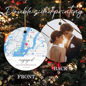 May include: A round, white, ceramic ornament with a map of New York City on the front. The map shows the location of the Empire State Building and the text "engaged" with the names "JACKSON" and "HAILEY" and the date "12/12/24". The back of the ornament features a photo of a couple in a romantic embrace.