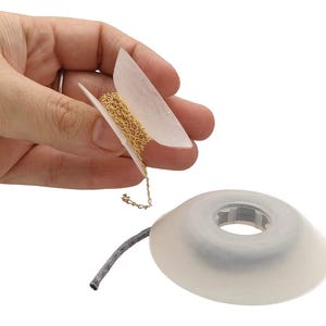 Puede incluir: Una mano sostiene una pequeña cadena dorada, parcialmente desenrollada de una tarjeta de papel blanco. Una bobina de cordón gris está unida a un dispensador de plástico blanco translúcido. La cadena es de color dorado brillante.