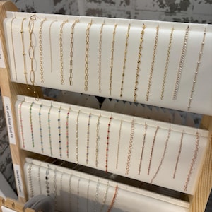 May include: A display of various necklaces, showcasing different chain styles and gemstone accents. The necklaces are arranged on a wooden rack with white trays. The chains are gold, silver, and rose gold in color.