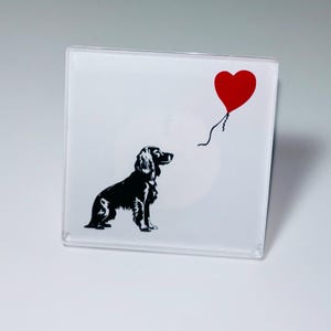 May include: Square, clear coaster with a black dog silhouette looking at a red heart-shaped balloon. The dog is detailed with fur and posture. The balloon is attached by a thin black string. The coaster has a glossy finish.
