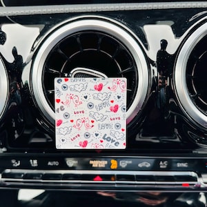 May include: A white car air freshener with a pink and red heart and bird design. The air freshener has the words "Love" and "Love Birds" written on it.