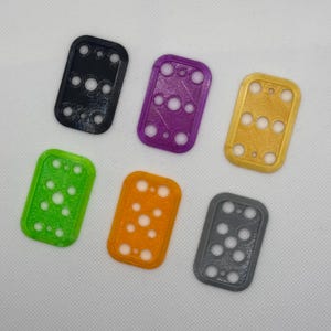 Puede incluir: Seis llaveros rectangulares de plástico en diferentes colores: negro, morado, dorado, verde, naranja y gris. Cada llavero tiene un patrón de agujeros diferente.