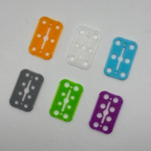 May include: Six plastic razor blade holders in different colours: orange, white, light blue, grey, green, and purple. Each holder has a rectangular shape with rounded corners and a slot in the centre for a razor blade.