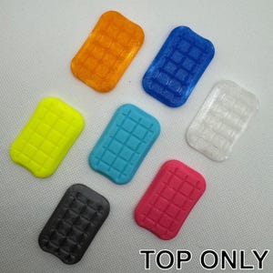 Puede incluir: Siete azulejos de plástico de diferentes colores con un patrón de cuadrícula. Los azulejos están dispuestos en semicírculo sobre una superficie blanca. Los colores son naranja, azul, transparente, amarillo, turquesa, rosa y negro.