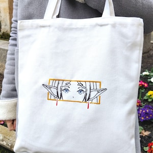 May include: White canvas tote bag featuring an embroidered image of an anime character's face with blue eyes and elf ears. The character's face is framed in a gold rectangle with red lines extending from the bottom.