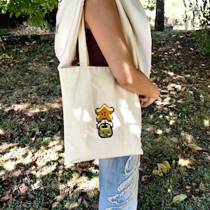 May include: A white canvas tote bag featuring a pixelated design of a yellow star and a green character. The bag is being carried by someone wearing a white hoodie and blue jeans.
