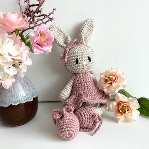 Pode incluir: Boneca de coelho de crochê feita à mão, vestindo um vestido rosa e sapatos combinando. O coelho tem orelhas compridas e uma faixa rosa. A boneca está ao lado de um vaso de flores cor-de-rosa e uma única flor.