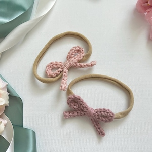 May include: Two elastic headbands featuring small, hand-knitted bows. One headband has a light pink bow, and the other has a dusty rose bow. Both headbands are made of a soft, tan-colored nylon material, designed to be stretchy and comfortable.