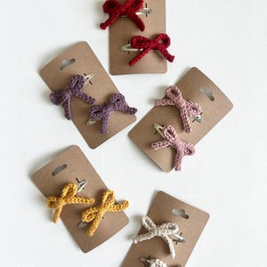 May include: Six sets of two crocheted hair bows on brown card stock. The bows are in red, pink, purple, yellow, and white.