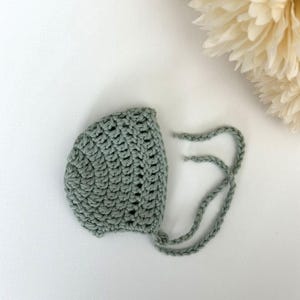 May include: A sage green crocheted baby bonnet with white ties.