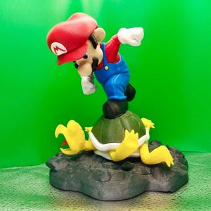 May include: A red and blue figurine of Mario, a video game character, stomping on a green and yellow Koopa Troopa. The figurine is made of plastic and is a 3D printed model.