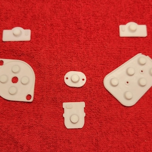 May include: White replacement buttons and directional pads for a game controller are arranged on a red surface. The components include various shapes, such as a D-pad, oval, and rectangular buttons, all with raised circular elements.