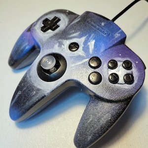 Könnte beinhalten: Ein individuell lackierter Nintendo 64-Controller mit einem Galaxie-Design. Der Controller verfügt über ein schwarzes Steuerkreuz, Tasten und einen Joystick. Der Körper ist mit einer Mischung aus Blau, Lila und Weiß bemalt, wodurch ein kosmischer Effekt entsteht.