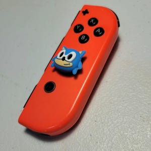 May include: A bright orange Nintendo Switch Joy-Con controller with a blue Sonic the Hedgehog character attached to the top.