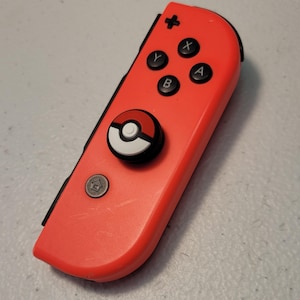 May include: A red Nintendo Switch Joy-Con controller with a black and white Pokeball design on the joystick.