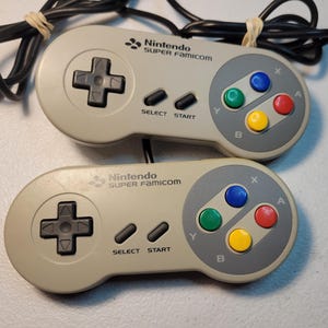 May include: Two gray Super Nintendo controllers with black cords. The controllers have the text "Nintendo SUPER FAMICOM" printed on them. The controllers have a directional pad, four face buttons, and two buttons labeled "SELECT" and "START".