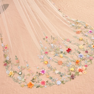 May include: A sheer, ivory-colored bridal veil with delicate floral embroidery. The veil features a cascade of colorful flowers in shades of red, yellow, pink, and blue, with green leaves. The flowers are clustered along the edges.