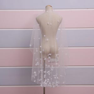 May include: A sheer white bridal veil adorned with delicate, three-dimensional white floral embellishments and scattered small beads. The veil is draped over a mannequin, showcasing its length and design. The background features horizontal stripes in pink, gray, and white.