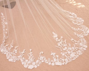 May include: A sheer, ivory-colored bridal veil with delicate floral lace detailing along the scalloped edge. The veil's fabric is lightweight and drapes gracefully, showcasing intricate white flower and leaf patterns.