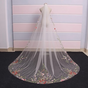 May include: A long, white bridal veil with delicate floral embroidery in red, pink, and green. The veil is displayed on a mannequin, showcasing its length and intricate design. The embroidery is concentrated along the edges.