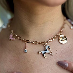 May include: Gold-toned charm necklace featuring a cowboy boot, a cowboy hat, a horse, and a dangling crystal. The necklace is worn around the neck. The chain is made of oval links.