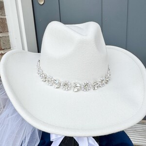 May include: A white cowboy hat with a rhinestone and pearl trim. The hat is sitting on a blue surface with a white veil.