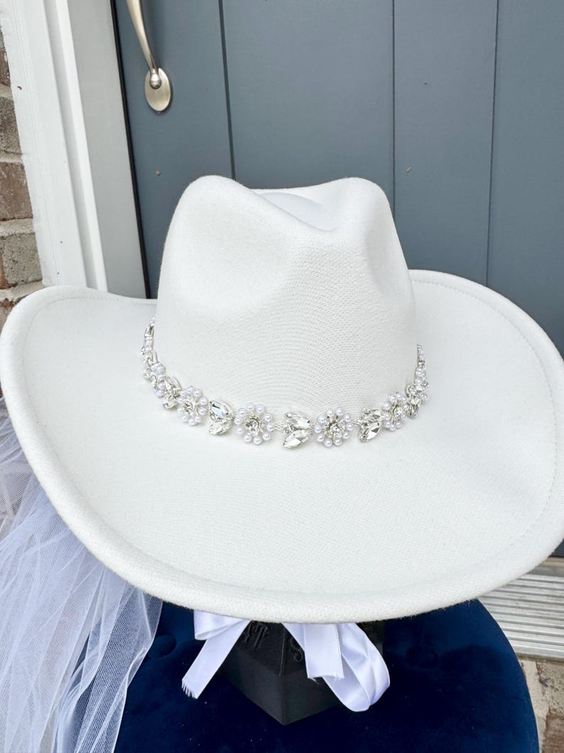 May include: A white cowboy hat with a rhinestone and pearl trim. The hat is sitting on a blue surface with a white veil.