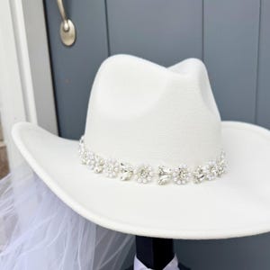 May include: White cowboy hat with a beaded trim of rhinestones and pearls. The hat is a bridal accessory.