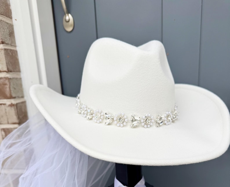 May include: White cowboy hat with a beaded trim of rhinestones and pearls. The hat is a bridal accessory.