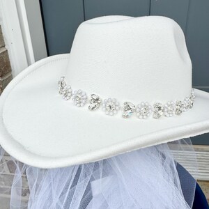 May include: A white cowboy hat with a beaded trim of pearls and rhinestones. The hat is paired with a white tulle veil.
