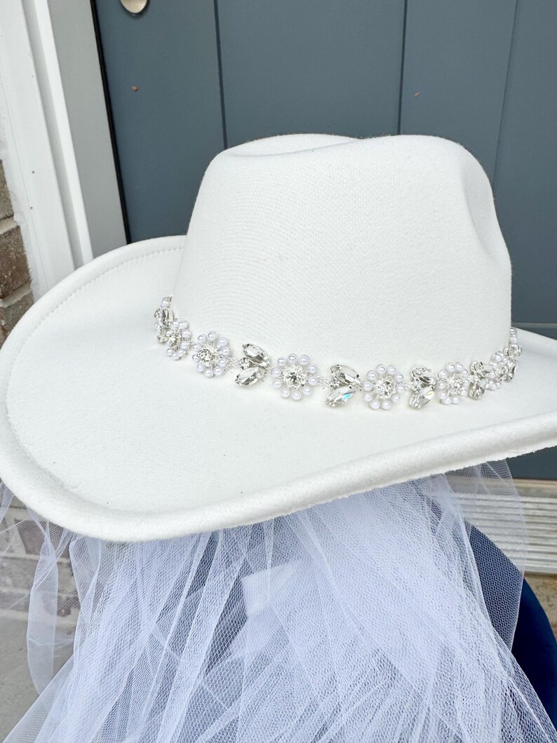 May include: A white cowboy hat with a beaded trim of pearls and rhinestones. The hat is paired with a white tulle veil.