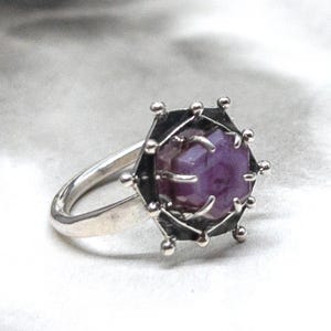 May include: A silver ring with a striking design. It features a faceted, deep purple gemstone set in a black, geometric frame. The ring's band is polished silver, and the gemstone is the main feature.