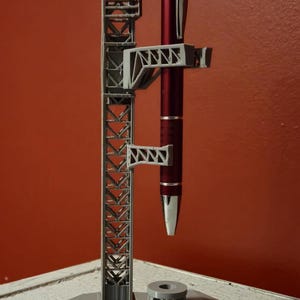 Puede incluir: Un portaplumas impreso en 3D con forma de torre de lanzamiento de cohetes. El portaplumas es gris y tiene un bolígrafo rojo dentro.