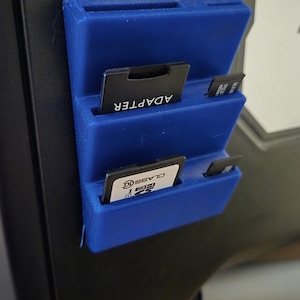 May include: A blue plastic storage case with three slots for memory cards. The top slot has a memory card labeled "ADAPTER", the middle slot has a memory card labeled "SDHC CLASS 10", and the bottom slot has a smaller memory card.