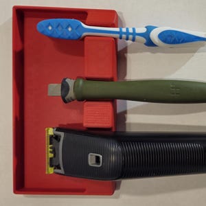 Puede incluir: Una bandeja de plástico roja con tres compartimentos contiene un cepillo de dientes azul y blanco, un cuchillo de uso general verde y negro y una afeitadora eléctrica negra.