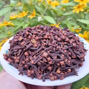 May include: A white plate piled high with a large quantity of dried, dark brown whole cloves. The cloves have a distinct nail-like shape with a rounded, textured head and a slender stem, showcasing their natural, earthy, and aromatic appearance.