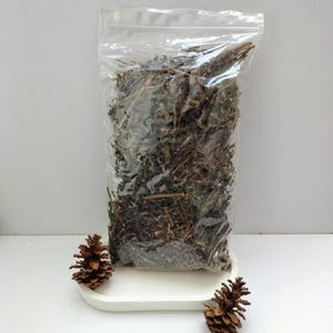 May include: A clear plastic resealable bag filled with a large quantity of dried brown plant stems and twigs. The bag is positioned on a white tray, flanked by two small brown pinecones on the left and right sides of the tray base.
