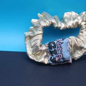 May include: A metallic gold fabric hair scrunchie featuring a light blue rectangular patch with the text BORN GIZ in black and pink lettering. The scrunchie has visible green stitching along the gathered fabric edges.