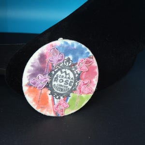 May include: A circular brooch featuring a watercolor-style background in shades of blue, purple, orange, and green. Centered is a black ornate frame containing the text "SERANA ROSE CIRCUS NIGHTMARE". Four pink embroidered flower-like shapes are attached.